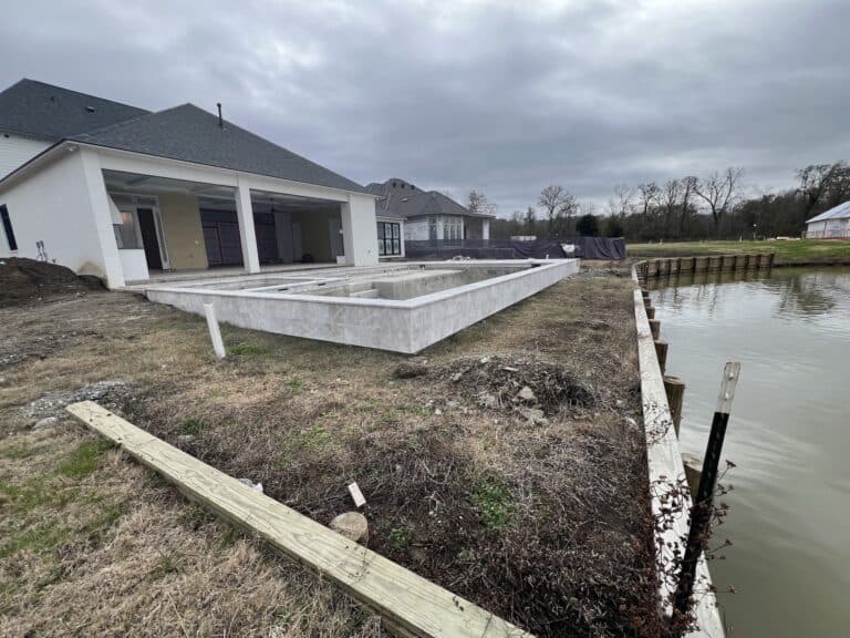 A concrete pool under construction behind a house near a canal, with overcast skies and grass surrounding the area.