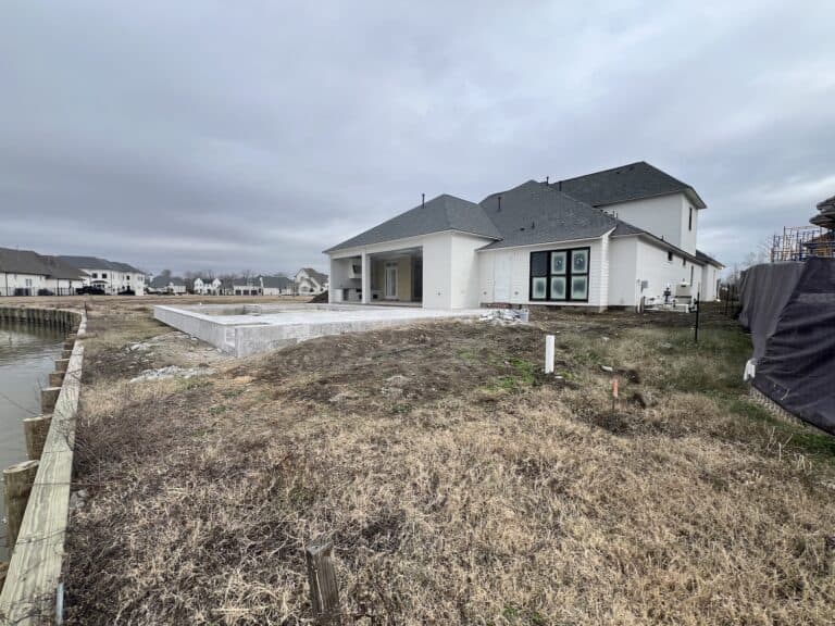 A house with gray shingles and white exterior stands next to a concrete foundation and a canal, with grass and cloudy skies.