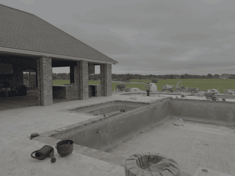 A partially constructed Lazy Waters backyard pool and hot tub with building materials scattered; one worker in an orange shirt stands nearby.