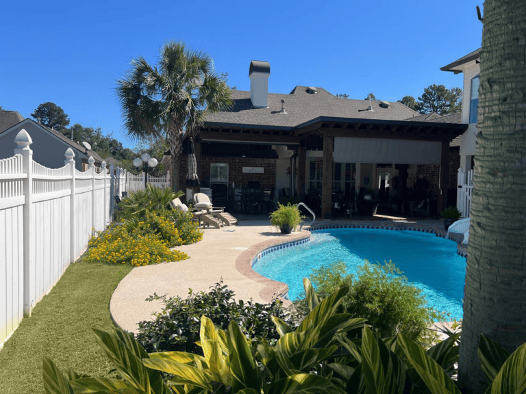 Backyard with a swimming pool, lounge chairs, palm trees, green plants, and a white fence beside a house under a clear blue sky.