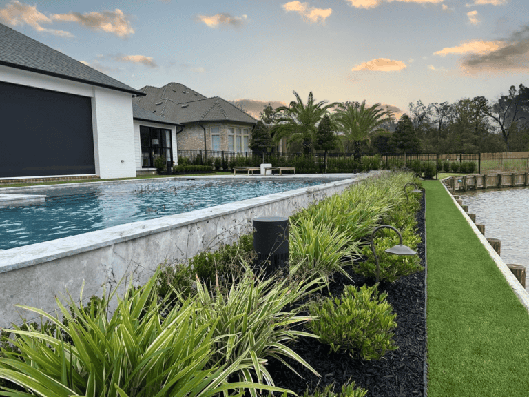 A backyard with a modern pool, landscaped plants, and Southern Moderne touches, overlooking a lake and sunset-lit homes in the distance.