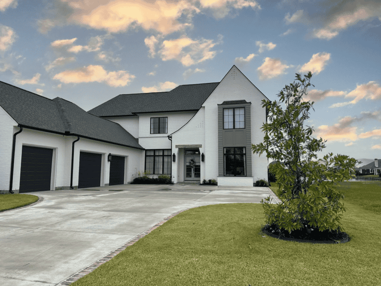 Southern Moderne two-story white house with black roof, three-car garage, large driveway, and a small tree under a partly cloudy sky.