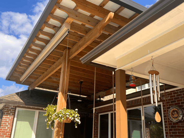 Wooden patio cover attached to a brick house, with hanging plants, wind chimes, and bird feeders visible under a sunny sky.