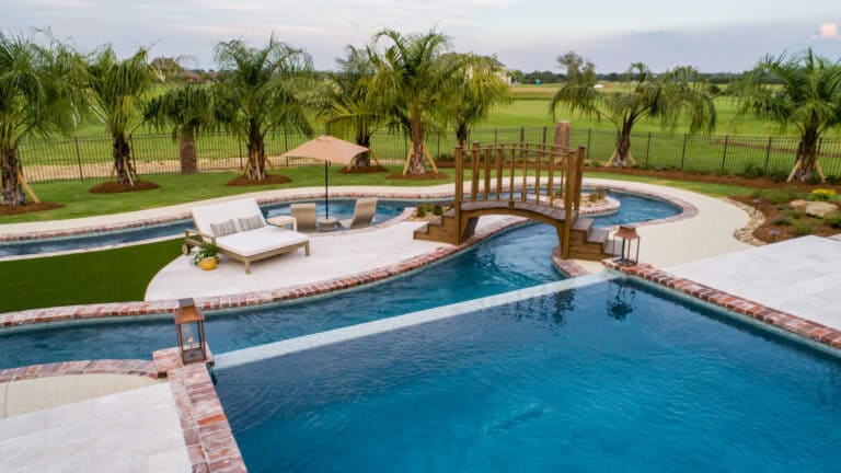 Rectangular resort-style swimming pool with a small wooden bridge, lounge chairs, palm trees, and grassy area in Lazy Waters' landscaped backyard.