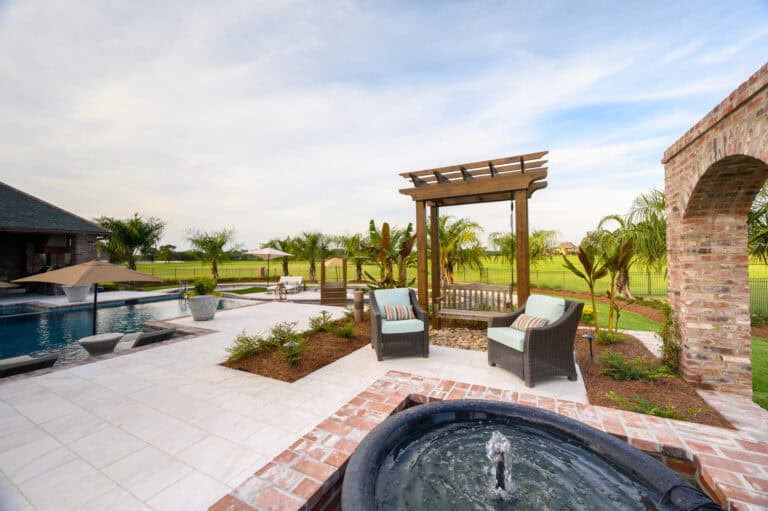 A landscaped backyard with a patio, wicker chairs, a pergola, Lazy Waters swimming pool, and a small fountain under partly cloudy skies.