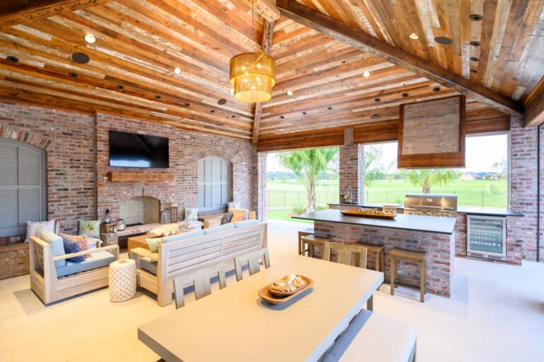 Spacious outdoor living area at Lazy Waters with wood ceiling, brick walls, lounge seating, TV, fireplace, dining table, and built-in appliances.