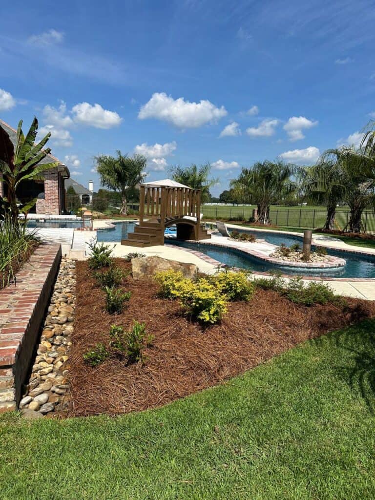 A landscaped backyard at Lazy Waters features a pool with a wooden bridge, palm trees, and mulched garden beds under a blue sky.