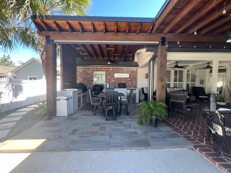 Outdoor patio with a stone floor, grill, dining table, covered wooden roof, and adjacent brick-floored seating area with chairs and fans.