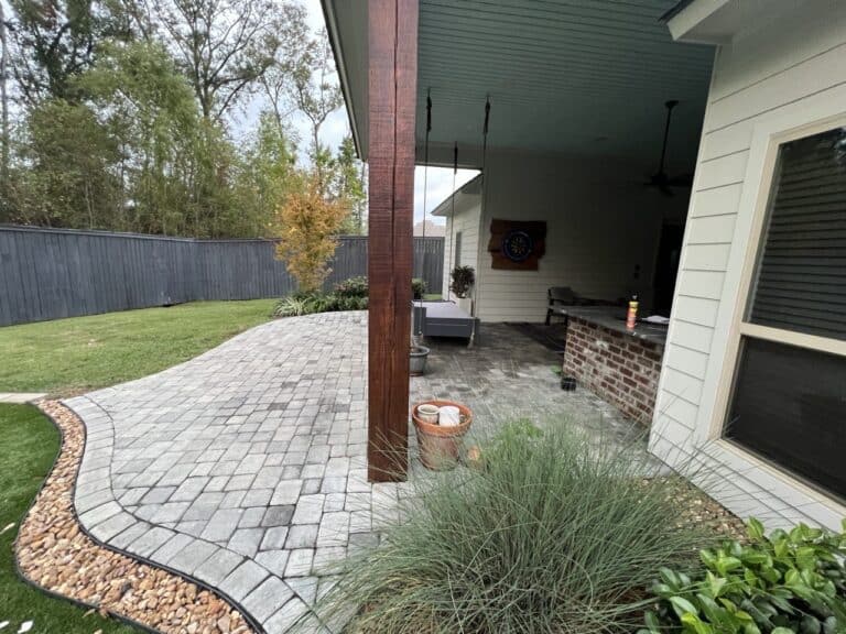Backyard patio with pavers, a covered seating area, and landscaped plants next to a lawn bordered by a wooden privacy fence.