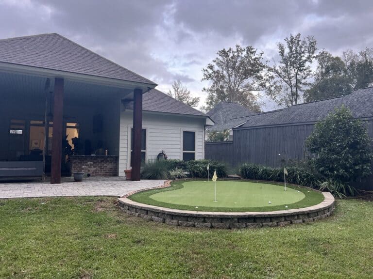 A backyard with a covered patio and a small artificial putting green surrounded by stone edging, under a cloudy sky.