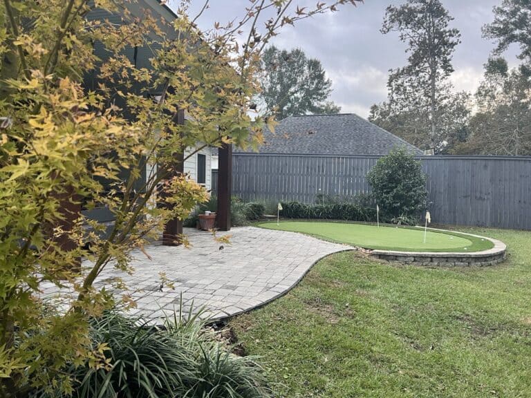 A backyard with a small putting green, paved patio, grass lawn, trees, and a wooden fence under a cloudy sky.