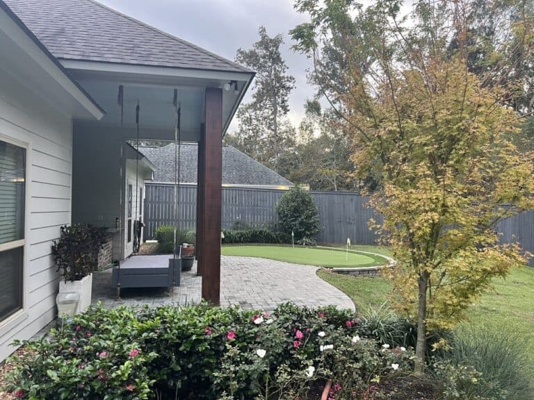 A backyard with a patio swing, paved area, small putting green, flower beds, a tree with fall foliage, and a wooden fence.