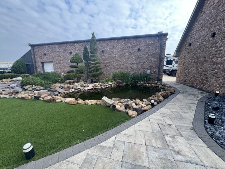 A landscaped yard at Pavillion Pointe features artificial grass, a rock-lined pond, brick buildings, a stone path, and an RV in the background.