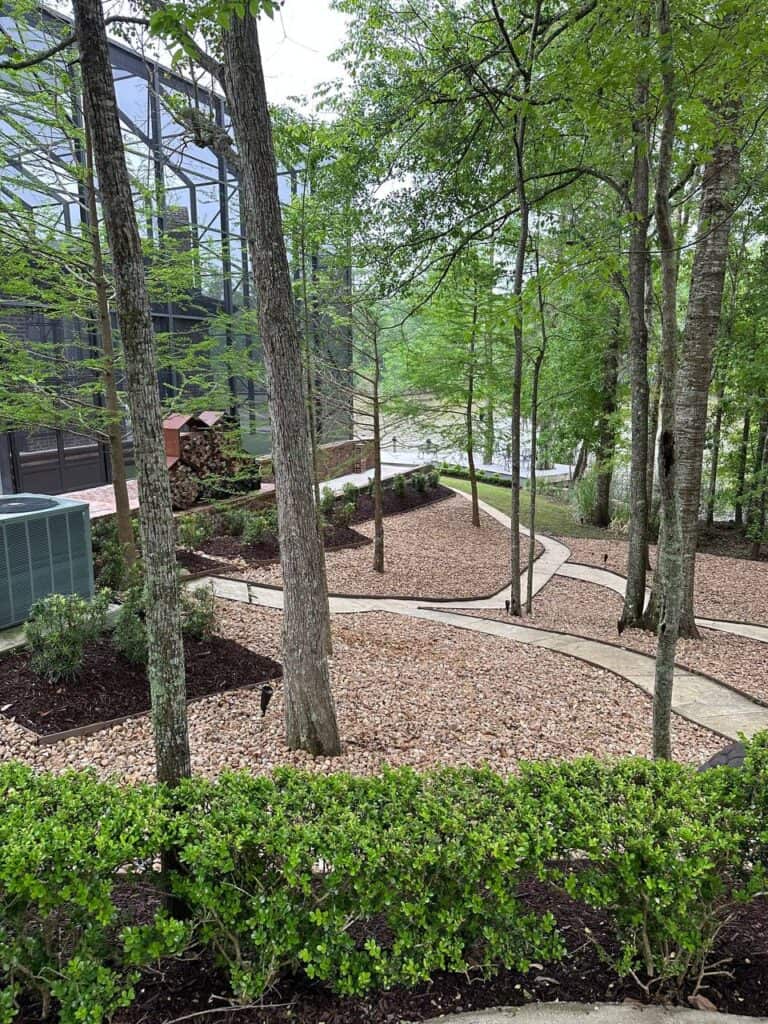 A gravel path winds through a landscaped area with trees, shrubs, and a large glass structure on the left.