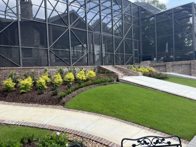 A neatly landscaped backyard with green grass, shrubs, a brick walkway, and a screened-in patio attached to a brick house.