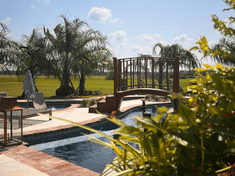 A swimming pool at Lazy Waters features a small arched wooden bridge, palm trees, and lounge chairs basking in the sunny day.