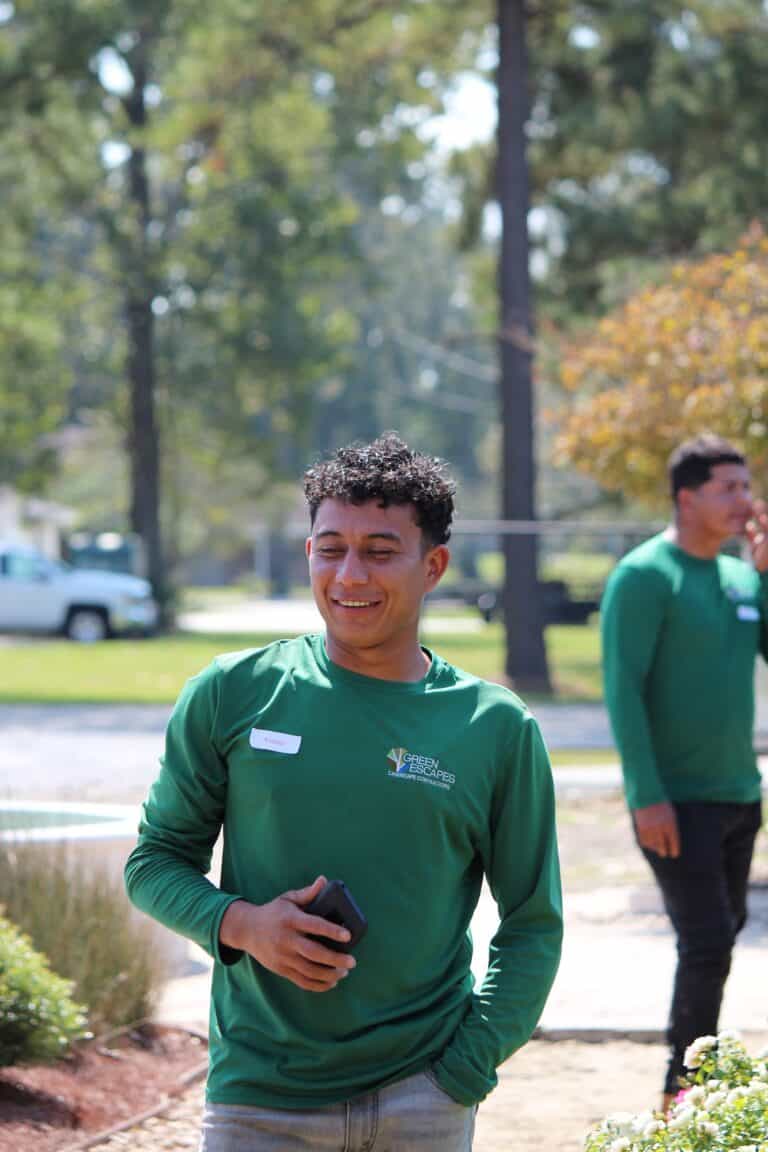 A man in a green long-sleeve shirt smiles outside, enjoying a Green Escapes Landscape with another person and trees in the background.