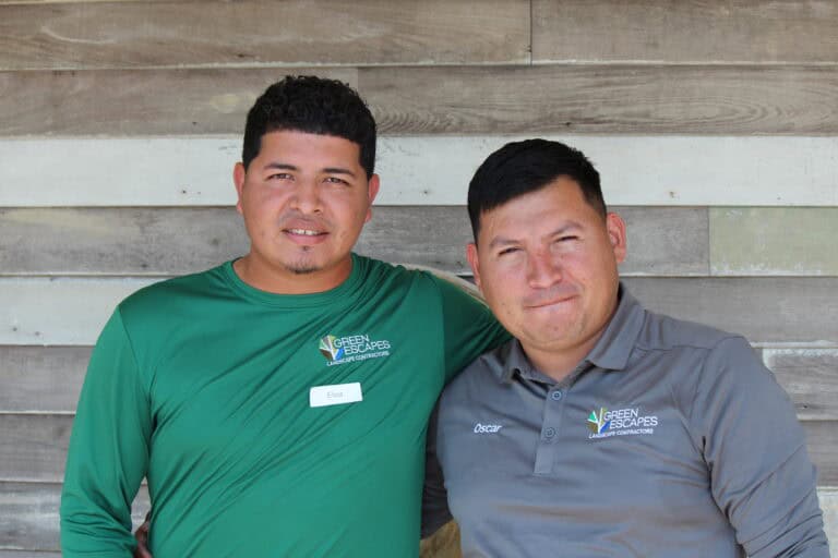Two men in “Green Escapes Landscape” shirts stand side by side before a wooden wall, one with his arm around the other.