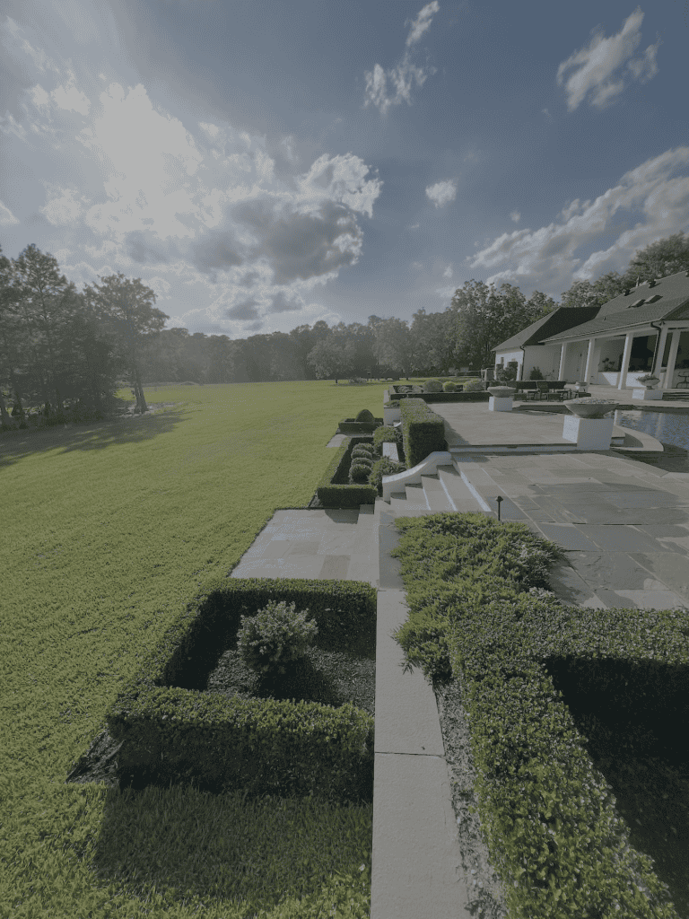 Landscaped backyard featured as an example portfolio piece, with trimmed bushes, patio, pool, lawn, and trees under a blue sky.