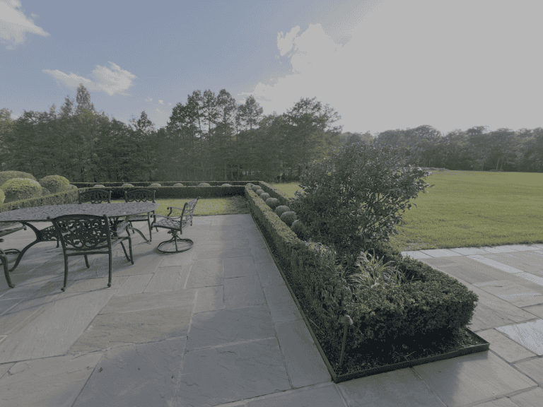 A stone patio with black metal table and chairs—an inviting portfolio piece—overlooks trimmed hedges, green lawn, and trees under a bright sky.