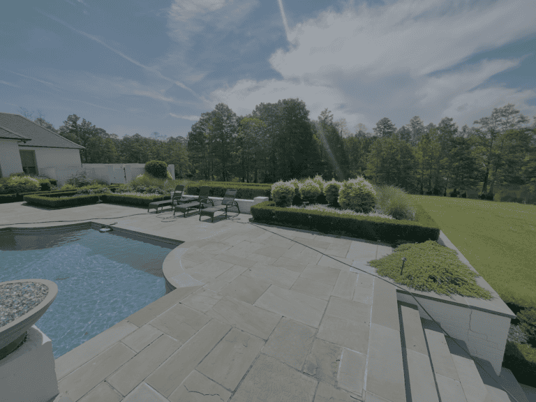Stone patio with lounge chairs by a pool, perfect for your portfolio, bordered by trimmed hedges, grass, trees, and blue sky.