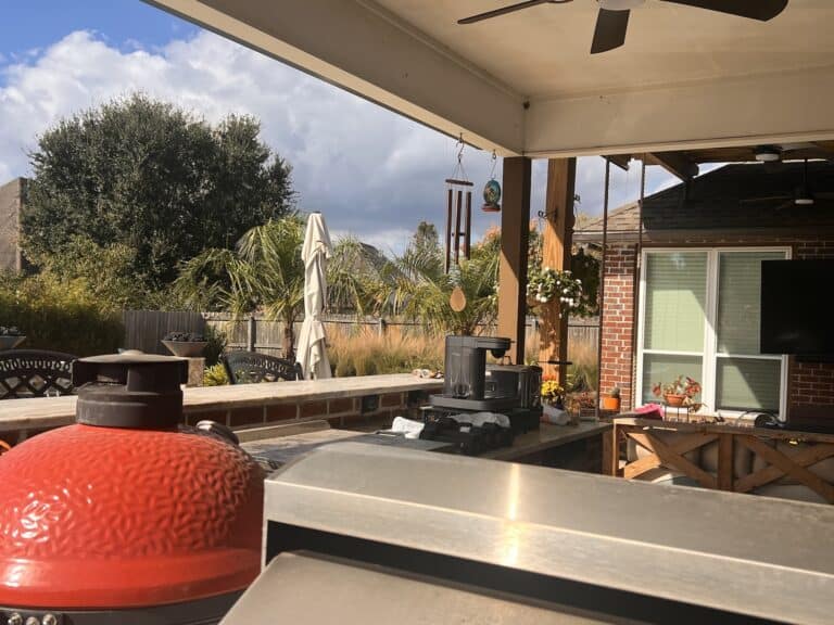Tropical Hideaway outdoor patio with a grill, red barbecue smoker, wind chimes, plants, and chairs under a covered area on a sunny day.