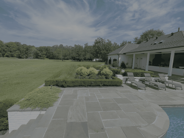 A stunning portfolio piece: a backyard with a stone patio, pool, lounge chairs, landscaped garden, lawn, and a white house under cloudy skies.
